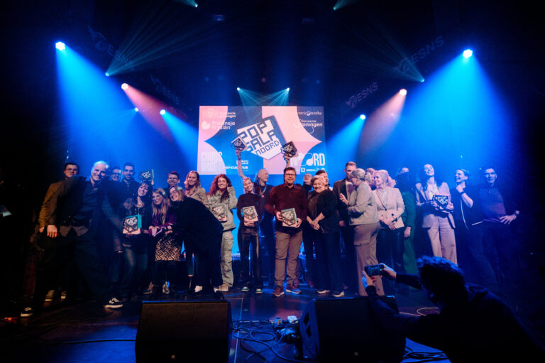 Groep mensen op een podium tijdens een evenement, met kleurrijke verlichting en een groot scherm op de achtergrond.