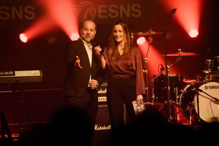 Twee sprekers staan op een podium met podiumverlichting, terwijl ze een evenement presenteren.
