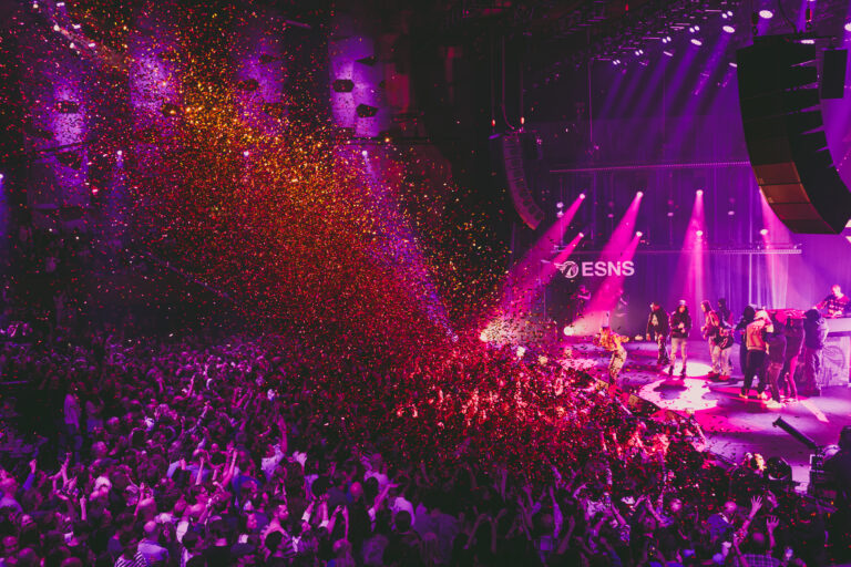 Een levendig concert met een enthousiast publiek dat wordt bedekt met kleurrijke confetti en heldere verlichting.