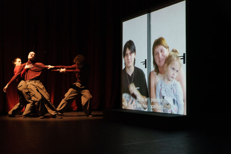 Een dansvoorstelling met performers die een emotionele interactie tonen naast een projectie van twee mensen met een kind.