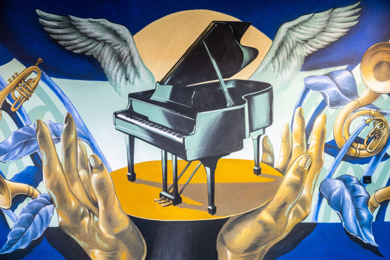 Een vleugelpiano met vleugels, omringd door grote gouden handen, tegen een fel oranje achtergrond en een blauwe lucht.
