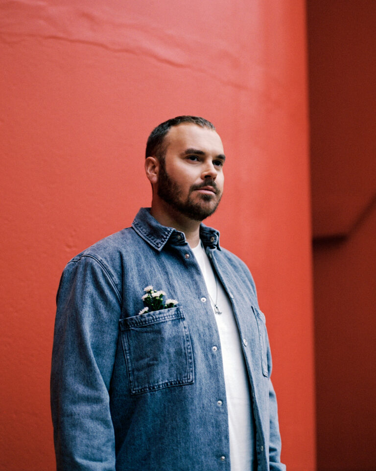 Een man met baard staat voor een felrode muur en draagt een denim jas.