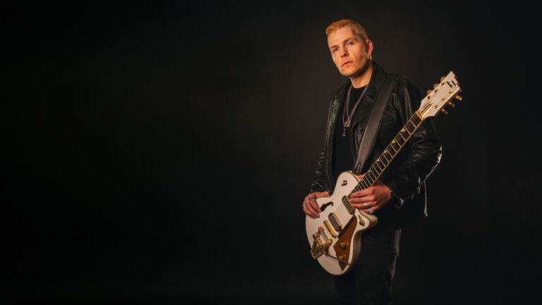 Een man speelt een witte elektrische gitaar tegen een donkere achtergrond.