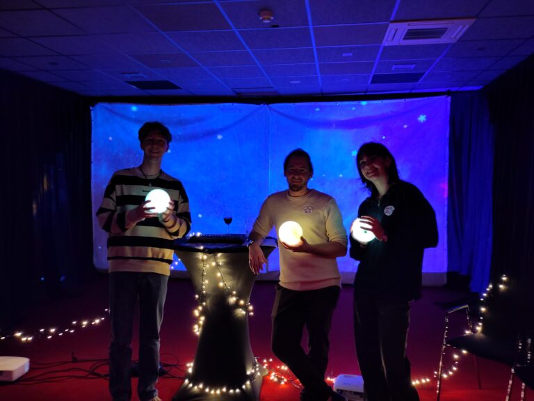 Drie mensen staan in een sfeervol verlichte ruimte, elk met een lichtgevende bol in hun hand, tegen een achtergrond van sterren en sfeervolle verlichting.