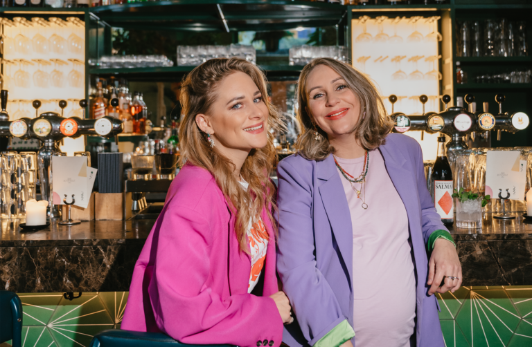 Twee lachende vrouwen staan samen achter een bar, waarbij de één een roze jas en de ander een lila jas draagt.