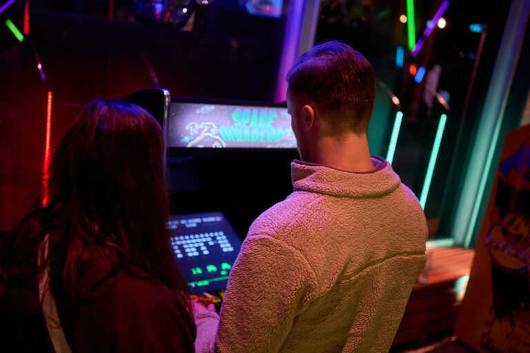 Twee mensen staan bij een retro arcade spelcomputer met kleurrijke verlichting op de achtergrond.