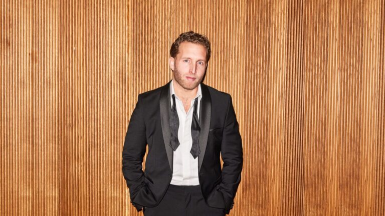 Een man met blond haar in een donker pak en wit overhemd staat voor een houten paneelachtergrond.