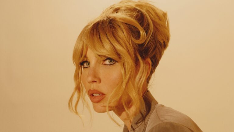 Portret van een vrouw met golvend, blond haar en een neutrale uitdrukking, gefotografeerd tegen een zachte, beige achtergrond.