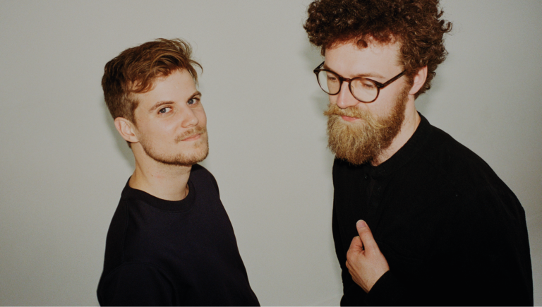 Twee mannen staan in een neutrale ruimte, met één man die naar de camera kijkt en de ander die iets naar hem toe gekeerd is.