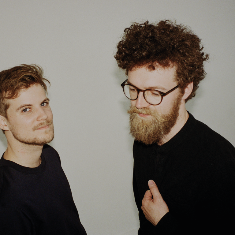 Twee mannen met verschillende haardrachten staan tegen een neutrale achtergrond, met de ene man met een volle baard en bril en de andere met korter haar.