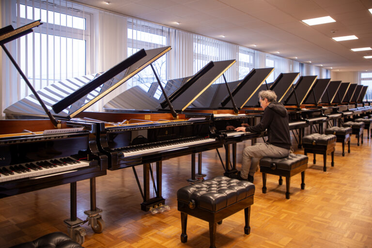 Meerdere zwarte piano's staan in een muziekruimte met houten vloer en ramen, met iemand op de achtergrond.