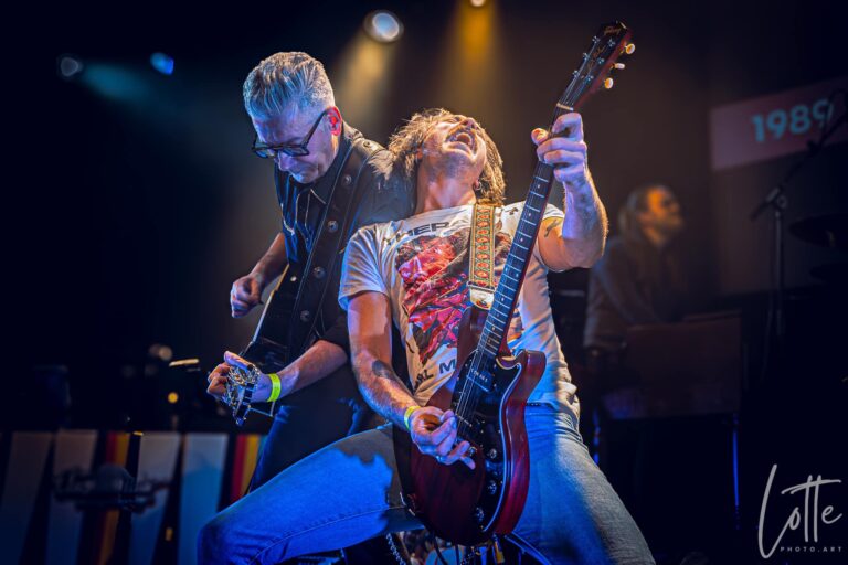 Een gitarist speelt een rode elektrische gitaar tijdens een live optreden, met een tweede muzikant op de achtergrond.