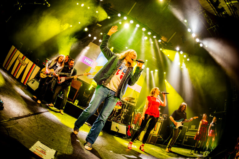 Een zanger met microfoon op een druk podium tijdens een concert, met een band op de achtergrond en felgekleurde lichten.