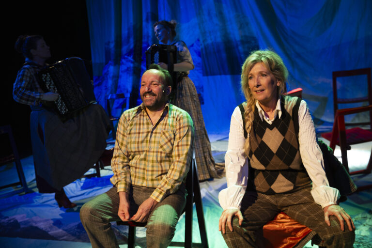 Twee mannen zitten op een podium tijdens een voorstelling, met blauwe verlichting op de achtergrond.