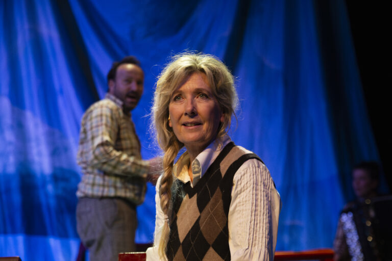 Een vrouw met blond haar draagt een argyle vest en staat op een podium voor een blauw gordijn, met een man op de achtergrond.