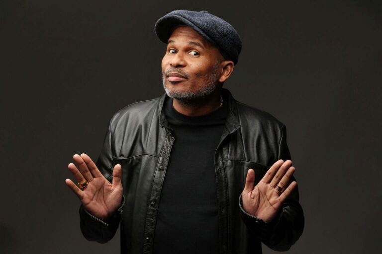 Een man in een zwarte leren jas en pet, met beide handen omhoog.