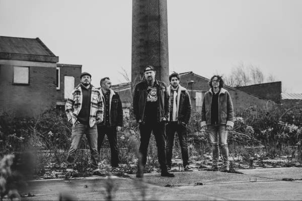 Vijf mensen staan in een rij op een vervallen straat vol puin, met een hoge schoorsteen op de achtergrond en de foto is zwart-wit.