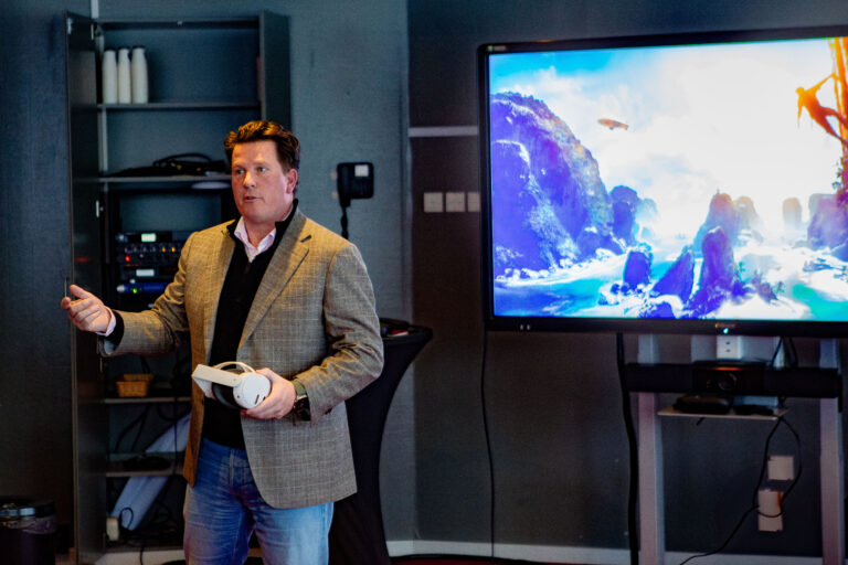 Een man in een grijs jasje houdt een witte bowlingbal vast tijdens een presentatie, met op de achtergrond een televisiescherm waarop een golffoto te zien is.