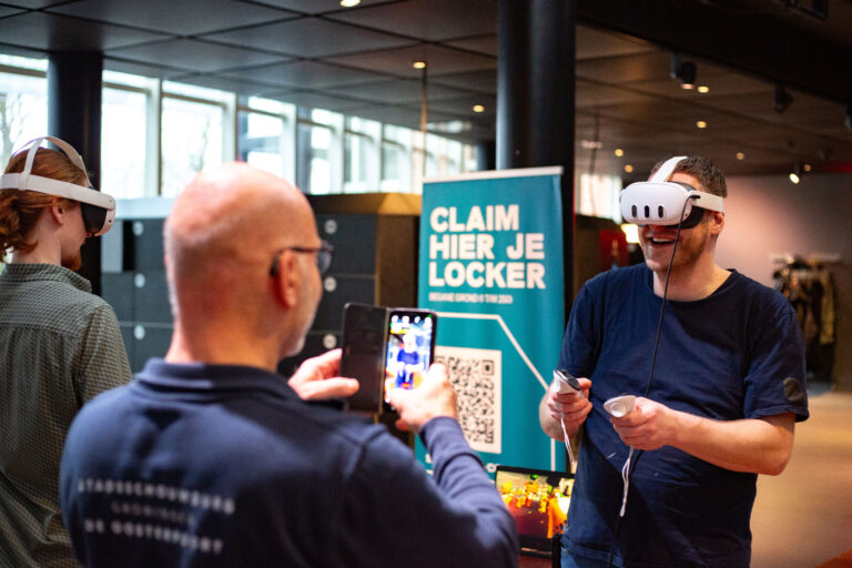 Man met kale kop houdt een telefoon vast naast een bord met de tekst “CLAIM HERE LOCKER”.