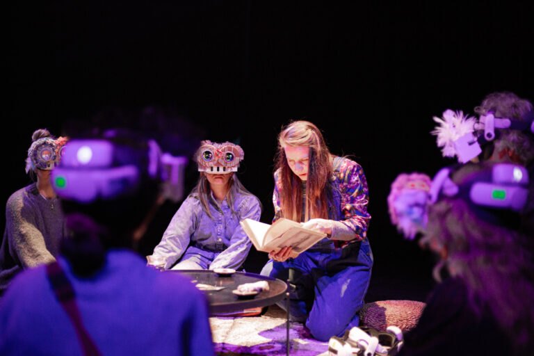 Een groep acteurs zit op een donker theaterpodium onder paars licht, terwijl een vrouw met lang haar een boek vasthoudt.