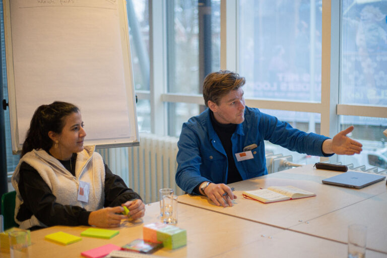 Twee personen zitten aan een tafel tijdens een vergadering met documenten en pennen, terwijl een flipover en ramen op de achtergrond te zien zijn.
