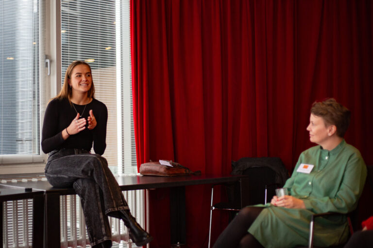 Twee mensen zitten op stoelen en praten in een ruimte met rode gordijnen op de achtergrond.