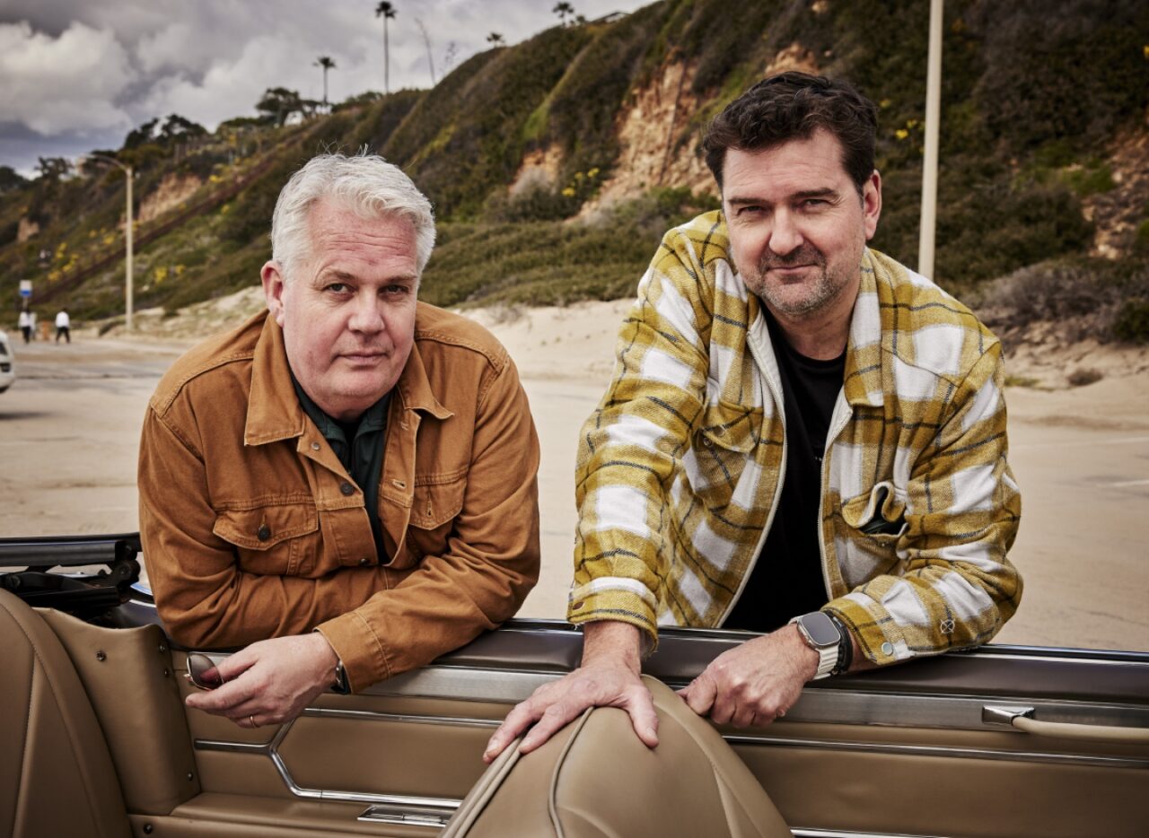 Twee mannen zitten naast elkaar aan een houten reling langs de kust, met kliffen op de achtergrond.