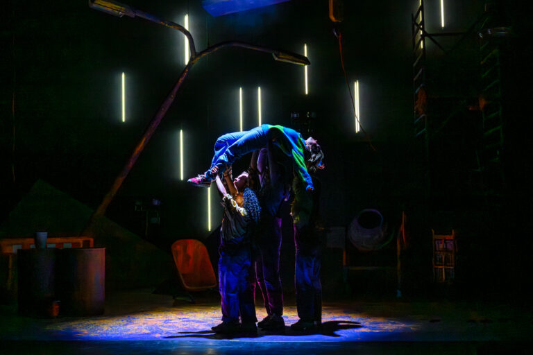 Twee acteurs op een podium, waarbij één acteur een andere ophoudt in een dramatische pose, omgeven door een sfeervolle verlichting.