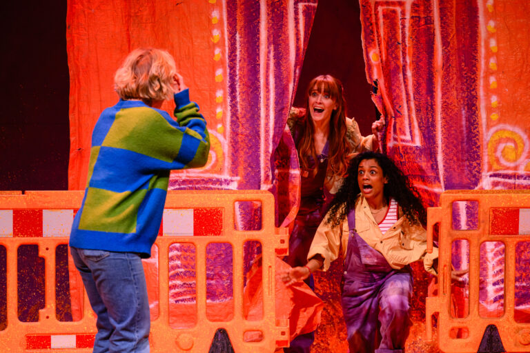 Drie acteurs op een podium, met een kleurvolle achtergrond, waarbij één acteur geschrokken achteruit deinst terwijl de andere twee naar voren komen.