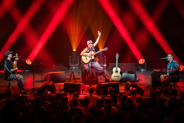 Een gitarist speelt gitaar op een podium onder fel rode lichten, met een publiek dat kijkt.