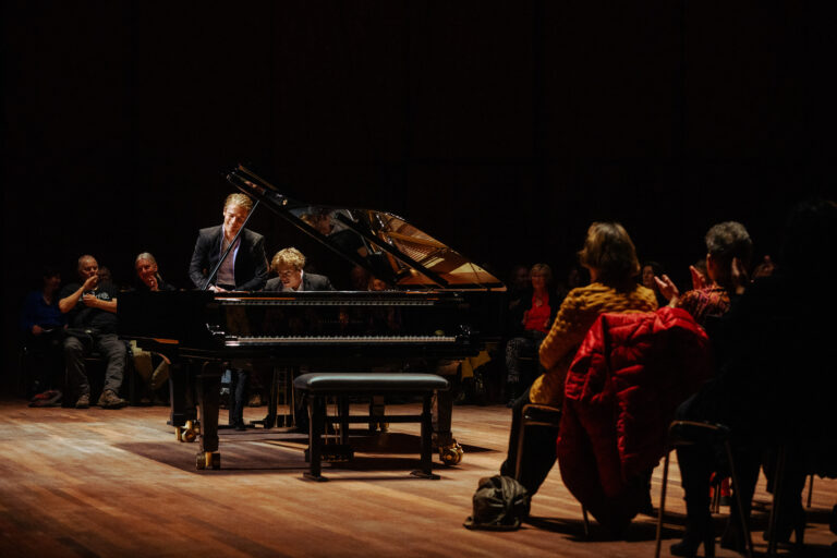 Een pianist speelt een vleugel op een donker podium, terwijl twee mensen in rode kleding erbij staan.