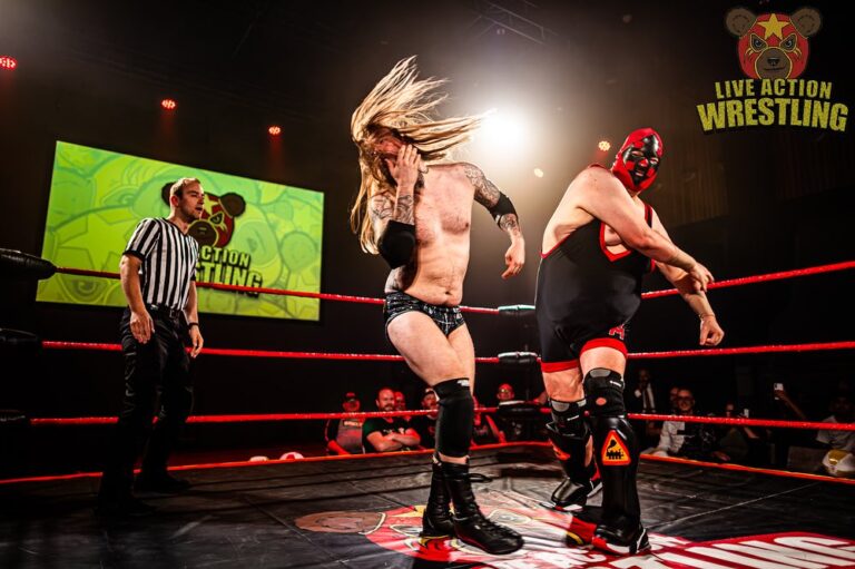 Een worstelaar met lang haar voert een actie uit in de ring, terwijl een scheidsrechter toekijkt.