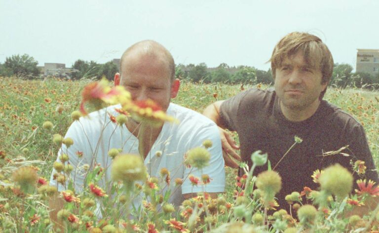 Twee mannen staan in een veld vol kleurrijke bloemen, met de voorste man in een lichtblauw T-shirt.
