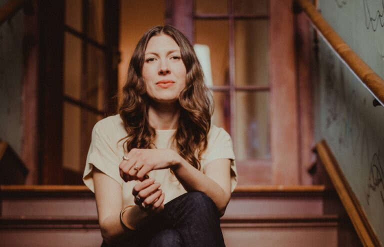 Een vrouw zit op een houten trap, haar handen gevouwen voor haar borst, terwijl ze naar de camera kijkt.