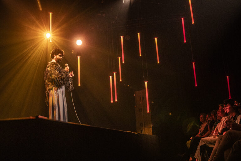 Een artiest op een podium tijdens een concert, met verticale rode lichtstralen op de achtergrond.
