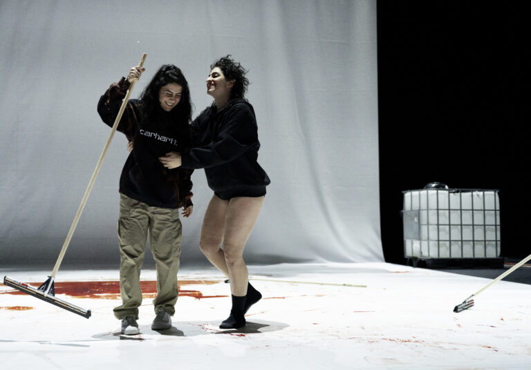 Twee dansers in casual kleding bewegen op een witte studiovloer tegen een grijze achtergrond.