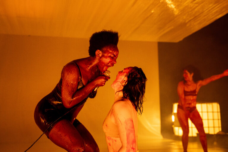 Een gespierde man in donkere kleding staat dicht bij een vrouw in een roze top in een warm verlichte ruimte; op de achtergrond is nog iemand te zien.