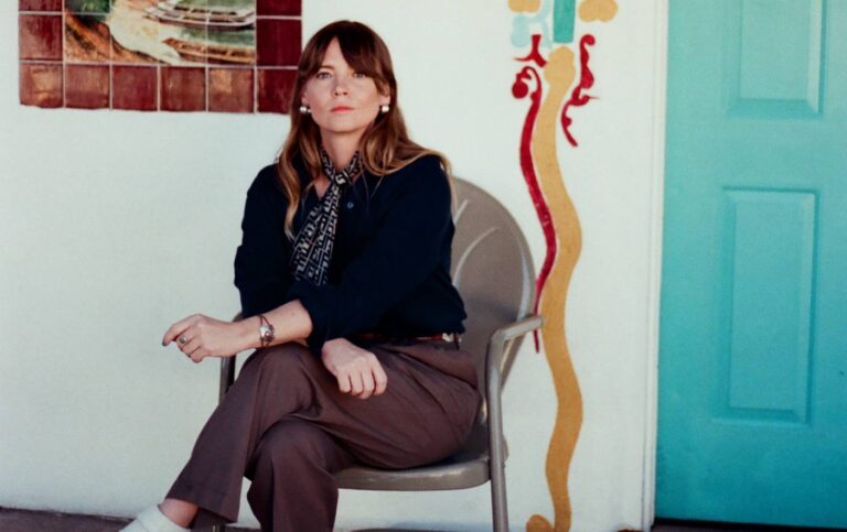 Een vrouw zit zittend op een stoel in een kamer met kleurrijke muurkunst op de achtergrond.