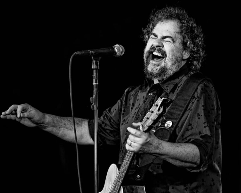 Een man zingt in een microfoon tijdens een live optreden, terwijl hij een gitaar vasthoudt.