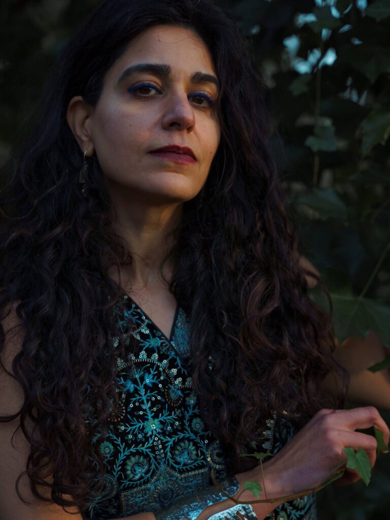 Portret van een vrouw met donker krullend haar, draag een blouse met turquoise patroon, buiten tegen een groene achtergrond.