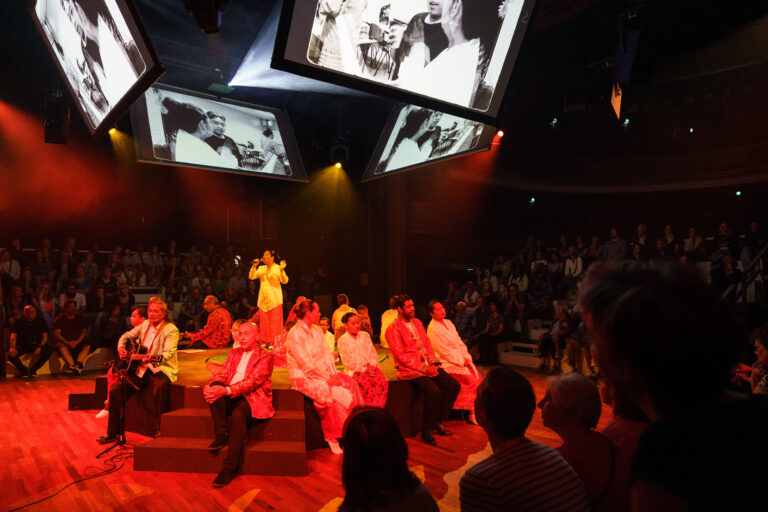 Een groep dansers in roze outfits voert een optreden uit op een verlicht podium in een theater, terwijl het publiek in de zaal kijkt.