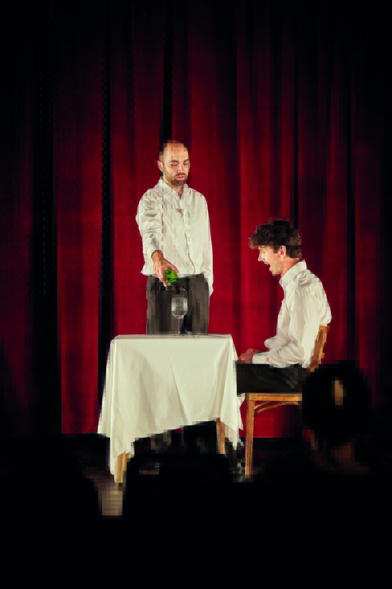Twee mannen op een podium rondom een tafel met een wit tafelkleed, waarbij één staat en de ander zit, tegen een rode gordijnachtergrond.