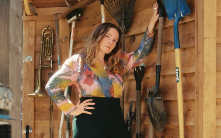 Een vrouw met een kleurrijke blouse en een zwarte rok poseert in een houten interieur, met één hand op haar heup.