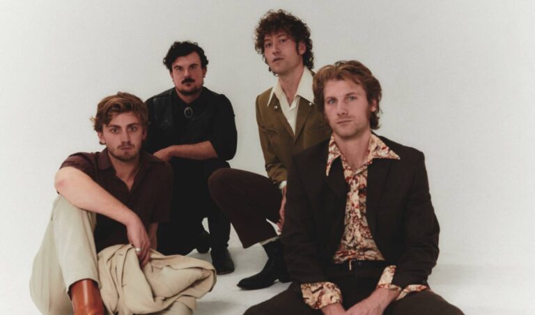 Vier mannen in seventies-stijl kleding poseren samen voor een witte achtergrond.