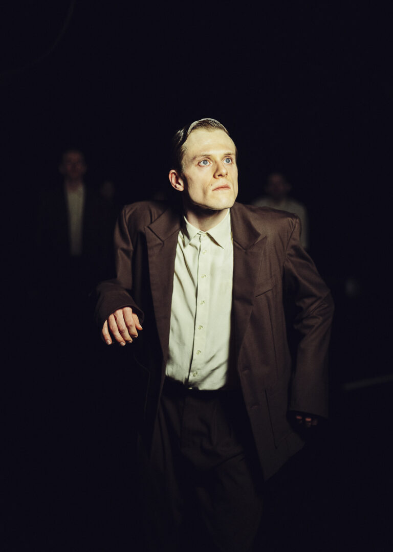 Een oudere man in een bruine jas en wit shirt loopt door een donkere ruimte, met licht op zijn gezicht.