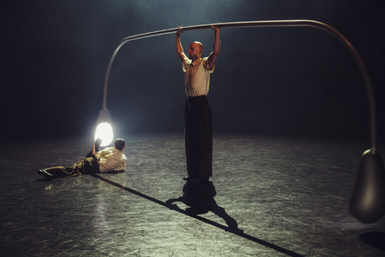 Twee mensen op een podium, waarbij één persoon een grote gebogen structuur vasthoudt terwijl de ander op de grond ligt.