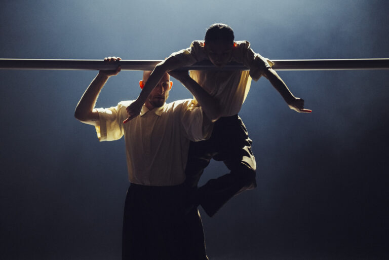 Twee personen tillen samen een halterstang boven hun hoofd tegen een donkere achtergrond.