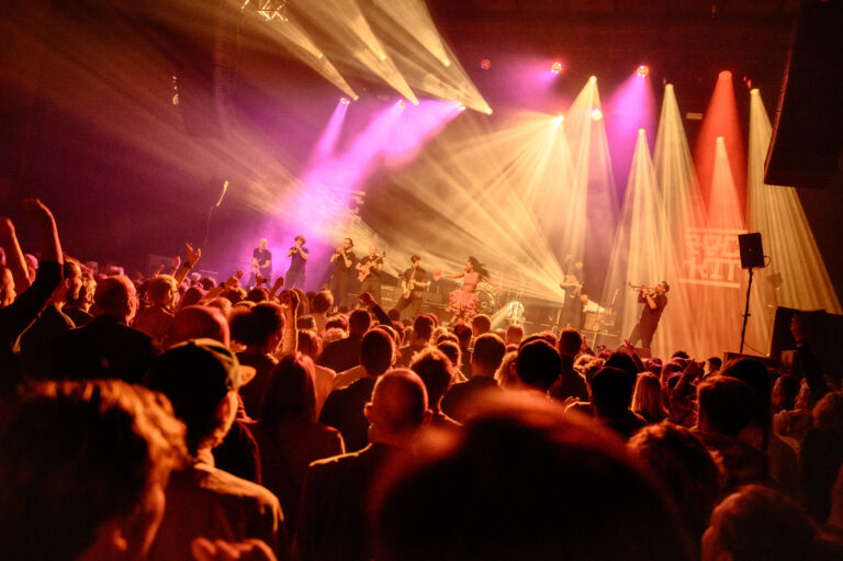 Drukke menigte op een concertvloer met felle, kleurrijke podiumverlichting.