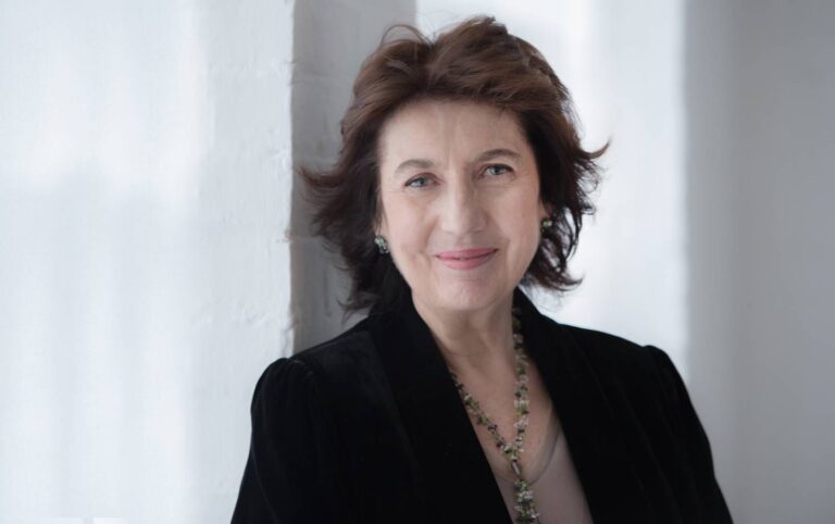 Portret van een vrouw met kort donker haar, die een zwarte blazer en een ketting draagt, glimlachend tegen een lichte achtergrond.