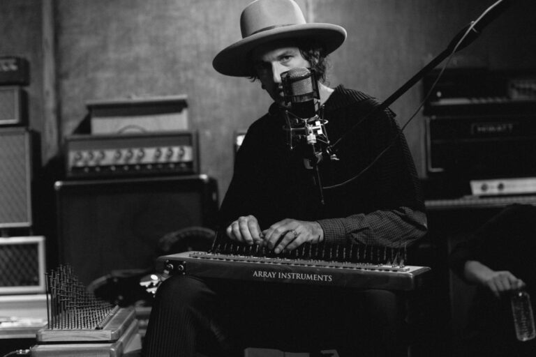 Een muzikant speelt op een gitaarachtig instrument terwijl hij in een studio zit, omringd door versterkers.